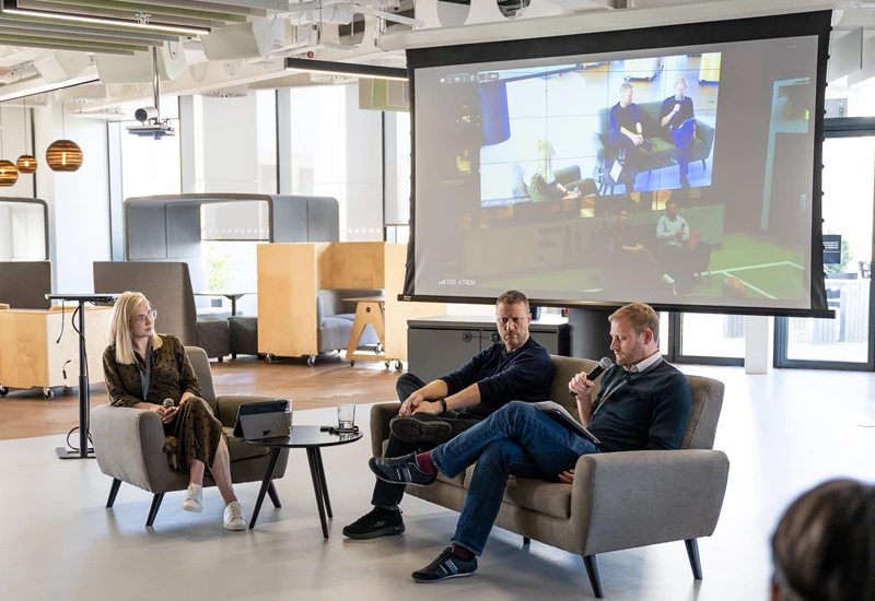 Three colleagues sit in office chairs, engaged in a discussion while a projector screen shows relevant content in the background